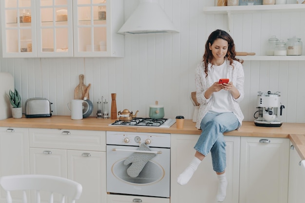 La casalinga femminile soddisfatta dei servizi di pulizia della casa si siede nella moderna cucina pulita con lo smartphone