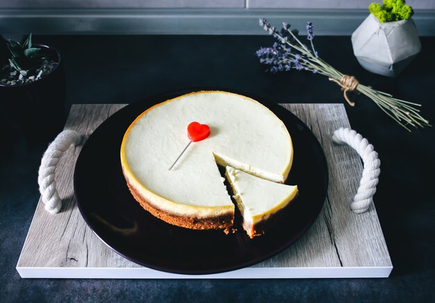 La classica cheesecake di New York fatta in casa giace su un piatto viola scuro su un vassoio di legno con manici in corda sul tavolo grigio della cucina. Lavanda e succulente sullo sfondo. Cuore rosso al regalo di San Valentino