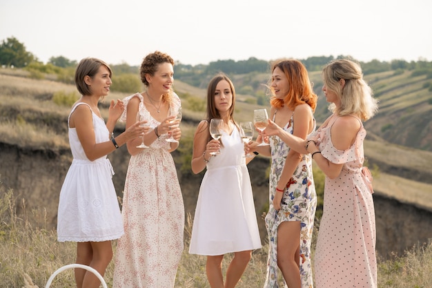 La compagnia di bellissime amiche che si divertono, bevono vino e si godono un picnic nel paesaggio collinare