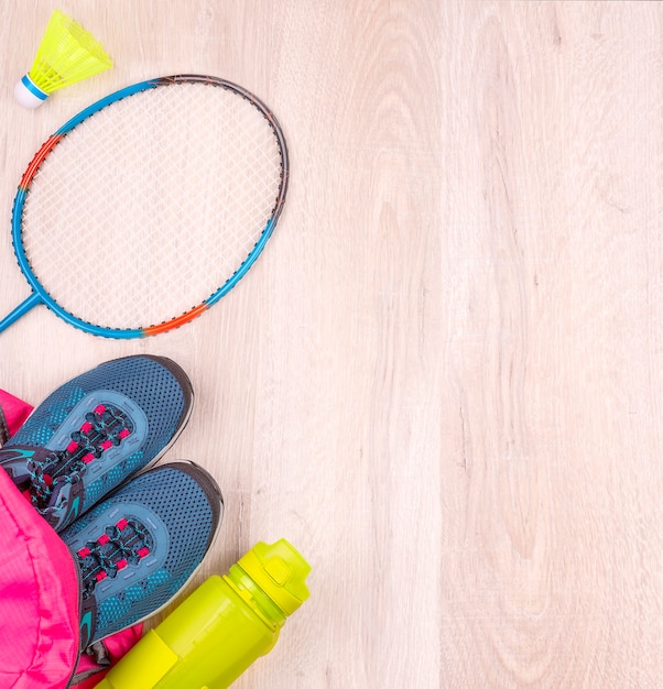 La coppia di scarpe da ginnastica da donna blu e bottiglia in zaino rosa, racchetta da badminton e volano su superficie di legno bianca