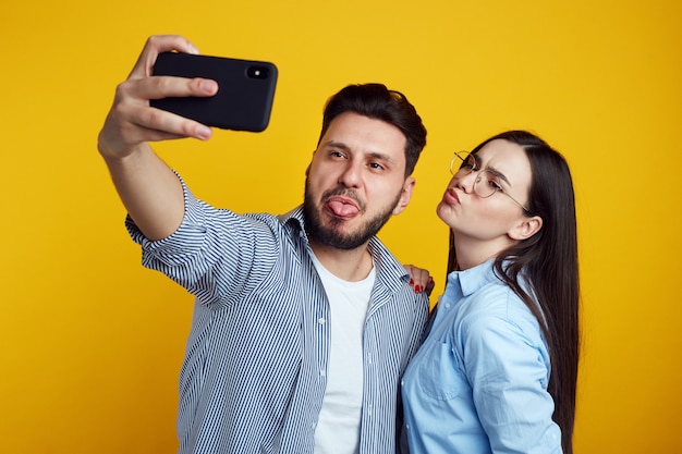 La coppia divertente fa la smorfia prende selfie sullo smartphone sopra la parete gialla