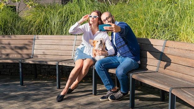 La coppia felice è seduta sulla panchina nel parco con un cagnolino e fa un selfie