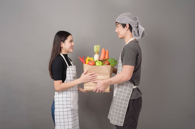 La coppia felice sta tenendo le verdure nella parete grigia del sacchetto della spesa
