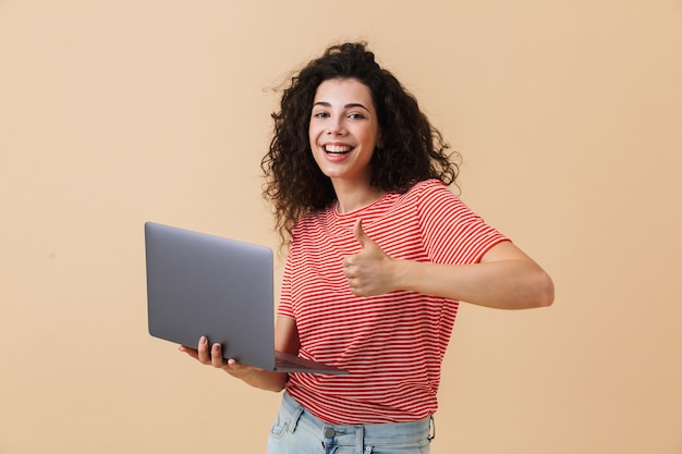 La donna abbastanza giovane riccia che per mezzo del computer portatile fa i pollici aumenta il gesto.