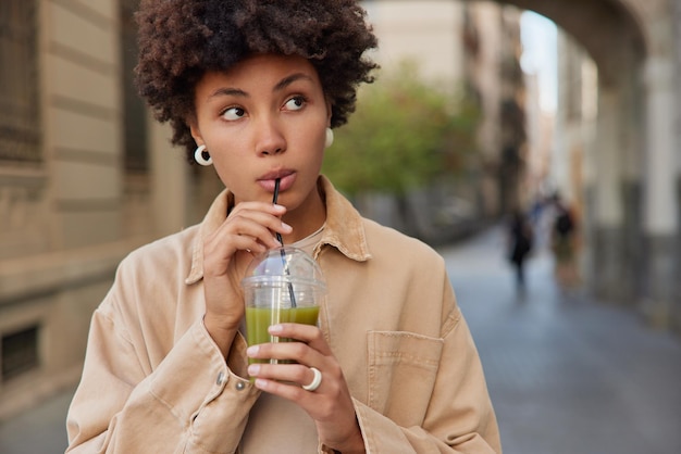 La donna abbastanza premurosa beve la bevanda disintossicante un cocktail verde biologico sano mantiene una corretta alimentazione vestita con abbigliamento da strada concentrato da qualche parte passeggiate nella città antica da solo Dieta e stile di vita