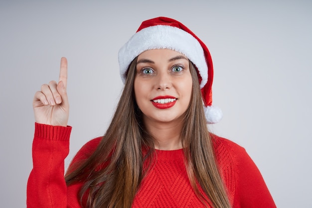 La donna adorabile in cappello della Santa, maglione rosso tiene il dito indice. Studio girato su sfondo grigio.