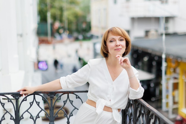 La donna adulta in abito bianco è in piedi sul balcone in città e sta guardando la fotocamera