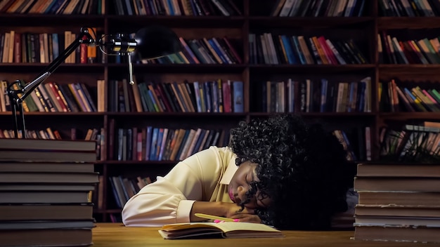 La donna afro-americana stanca di prepararsi ai test dorme vicino a un quaderno di carta aperto e una pila di libri sul tavolo contro il primo piano degli scaffali