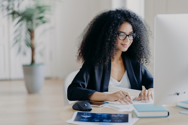 La donna afro indipendente lavora a distanza, scrive informazioni, si concentra sullo schermo del computer con espressione felice