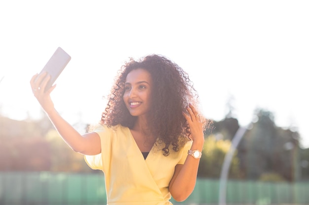 La donna afro sorridente con i capelli ricci che si fa un selfie durante il tramonto include spazio per la copia e il testo