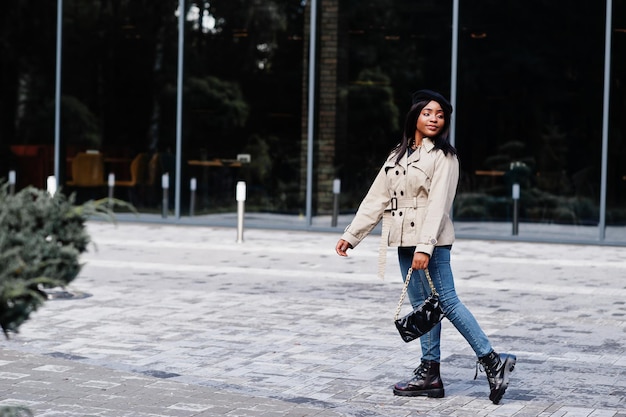 La donna afroamericana alla moda indossa il berretto e il cappotto con la borsa in posa all'aperto.