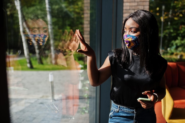 La donna afroamericana alla moda indossa una maschera per il viso fatta a mano contro la finestra