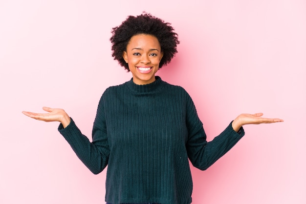 La donna afroamericana di mezza età su uno sfondo rosa isolato fa la scala con le braccia, si sente felice e fiduciosa.