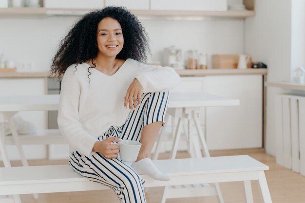 La donna afroamericana felice tiene la tazza di bevanda calda si appoggia al ginocchio indossa un maglione bianco elegante e pantaloni a righe