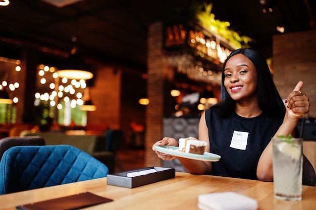 La donna afroamericana femminista alla moda indossa una maglietta nera e pantaloncini, posata al ristorante, mangia una torta di formaggio.