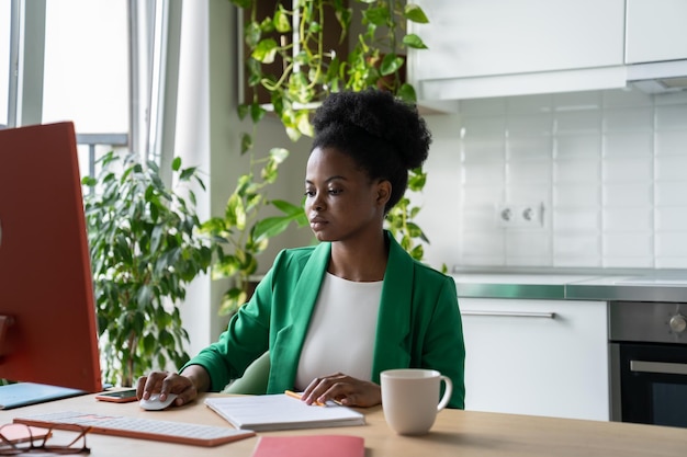 La donna afroamericana focalizzata libera professionista si siede al tavolo con il computer e lavora a distanza a casa