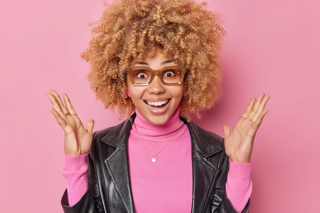 La donna allegra iperemotiva reagisce alle notizie fantastiche tiene le mani alzate sorride ampiamente indossa occhiali dolcevita casual e giacca di pelle isolata su sfondo rosa. Concetto di reazioni umane