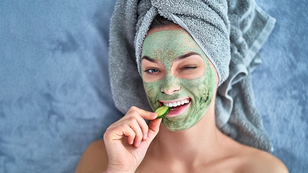 La donna ammiccante allegra felice con la maschera di pulizia verde dell'argilla e la fetta del cetriolo in asciugamano di bagno sta riposando e rilassandosi durante il giorno della stazione termale a casa. Cura della pelle del viso