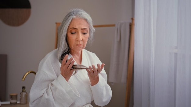 La donna anziana che tocca i capelli lisci lunghi grigi si preoccupa per il problema dell'invecchiamento della calvizie debole dell'attaccatura dei capelli persa