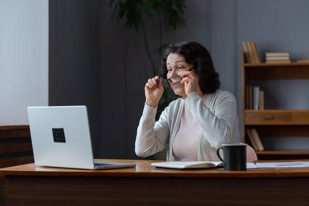 La donna anziana di mezza età felice si siede con il computer portatile che parla durante la videochiamata con la famiglia degli amici che ride matur