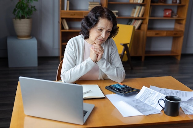 La donna anziana di mezza età si siede con il computer portatile e il documento cartaceo