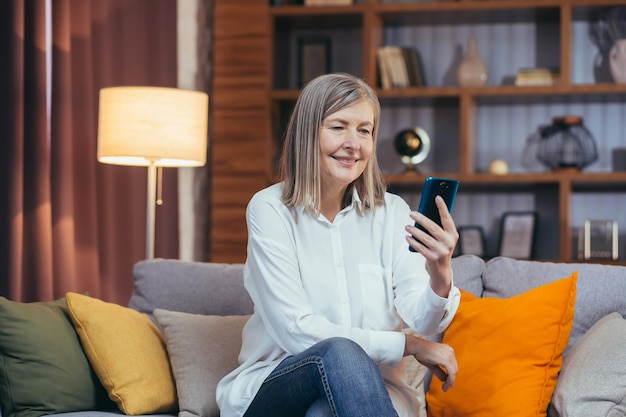 La donna anziana felice usa il telefono per la comunicazione online seduta sul divano a casa la sera e sorridente