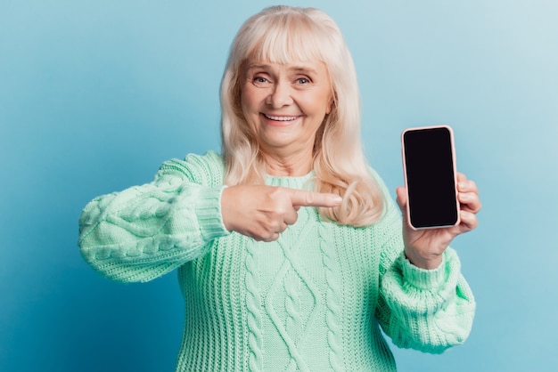 La donna anziana positiva tiene il touch screen del dito del punto dello smartphone isolato su fondo blu