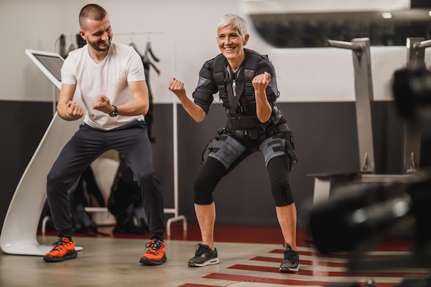 La donna anziana sorridente sta facendo EMS che lavora con il personal trainer in palestra.