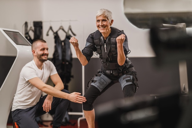 La donna anziana sorridente sta facendo EMS che lavora con il personal trainer in palestra.