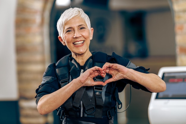 La donna anziana sorridente sta mostrando la forma del cuore sull'allenamento EMS in palestra. Guardando la fotocamera.