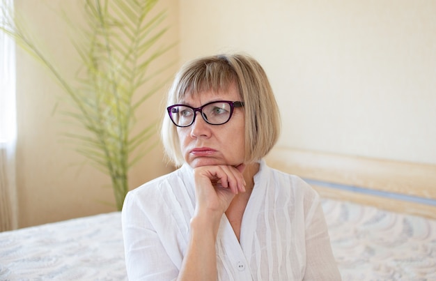 La donna anziana triste con i capelli bianchi di occhiali è triste nella sua casa in camera da letto. Concetto di solitudine, desiderio, tristezza