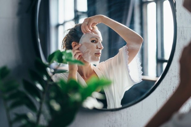 La donna applica la maschera in foglio al viso guardandosi allo specchio