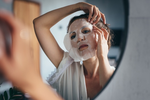 La donna applica la maschera in tessuto sul viso in bagno.