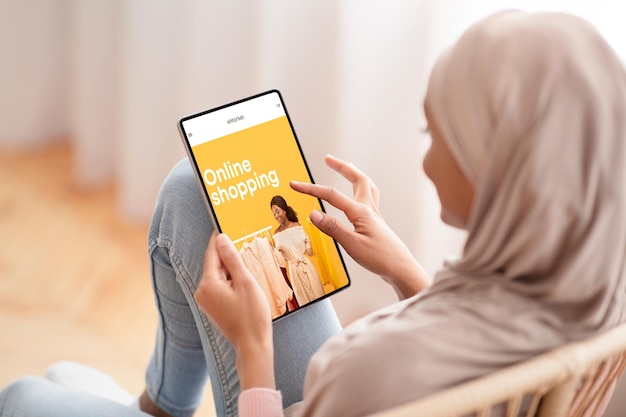 La donna araba millenaria nera in hijab che usa il tablet con il negozio di moda sullo schermo sceglie i vestiti a