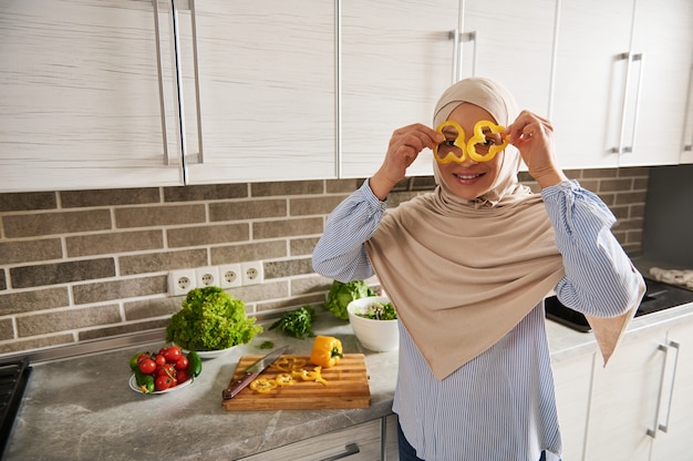 La donna araba musulmana in hijab guarda attraverso le fette di peperone giallo, gustando un'insalata vegana di preparazione in cucina