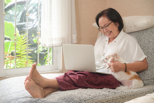 La donna asiatica anziana che si siede su un sofà sta usando un computer portatile. Lei sorrise felicemente, il cane chihuahua era seduto di lato.