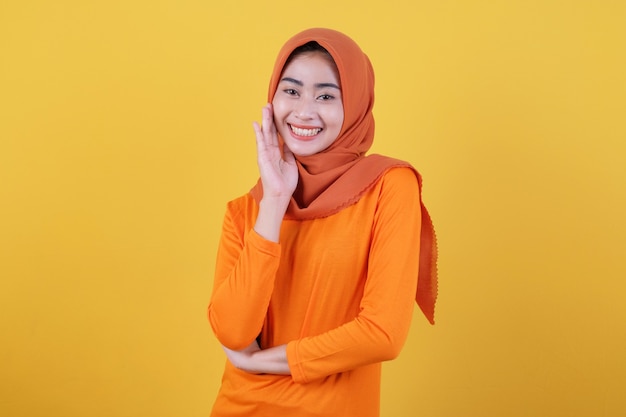 La donna asiatica attraente allegra dimostra lo spazio della copia sulla parete gialla in bianco, ha un'espressione amichevole felice, vestita casualmente che indossa l'hijab, posa al coperto