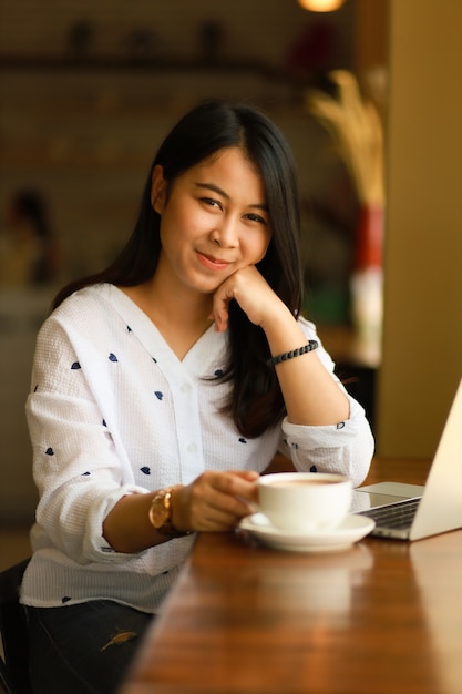 La donna asiatica che usando il computer portatile funziona e beve il caffè in caffè