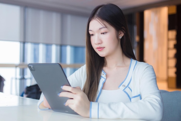 La donna asiatica con i capelli lunghi in camicia bianca tiene il tablet sorridente mentre si siede sulla sedia