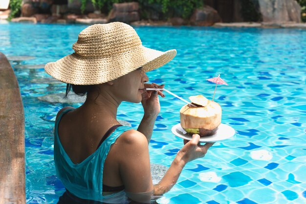 La donna asiatica con il cappello che mangia la noce di cocco beve nella piscina