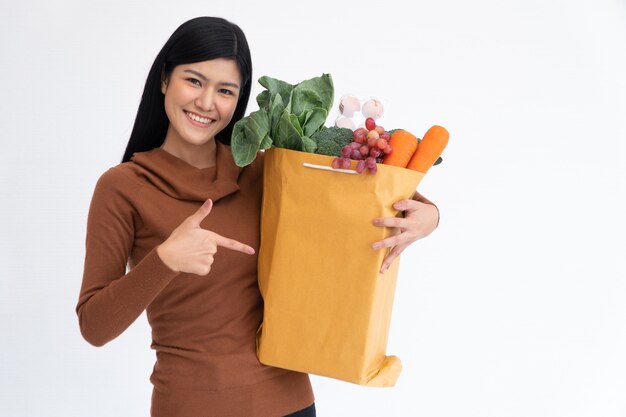 La donna asiatica felice che sorride e punta il dito verso un sacco di carta e porta un sacchetto della spesa