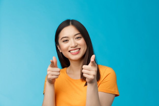 La donna asiatica moderna e amichevole allegra mostra gioiosamente le pistole con le dita, puntando la telecamera, sorridendo ampiamente, congratulandosi con l'amico buon lavoro, incoraggiandoti a continuare a lavorare, in piedi sfondo blu.