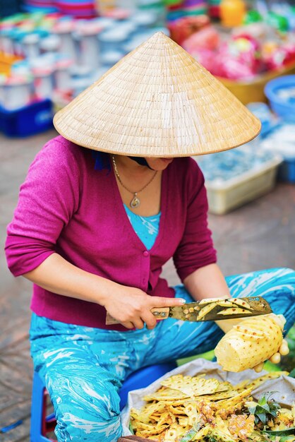 La donna asiatica nel tradizionale cappello vietnamita sta tagliando l'ananas nel mercato di strada a Can Tho, Vietnam