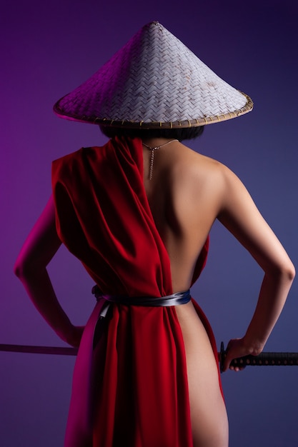 La donna asiatica snella nuda in un mantello rosso e un cappello asiatico con una katana in mano immagine di un samurai su uno sfondo al neon vista dal retro