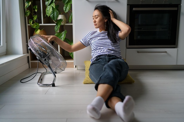 La donna asiatica soddisfatta soffiata da un ventilatore elettrico si siede sul pavimento e si gode il vento fresco dal ventilatore dell'aria in calore