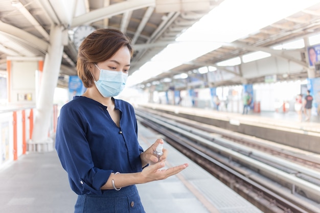 La donna asiatica sta aspettando il treno alla stazione. Spruzza alcol per pulirsi la mano e indossa una maschera medica come assistenza sanitaria e nuovo stile di vita normale.