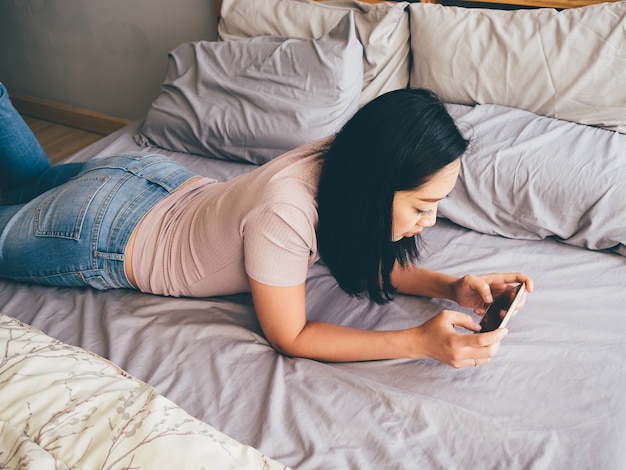 La donna asiatica sta giocando il gioco dello smartphone sul suo letto.