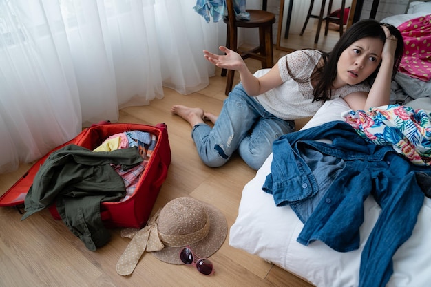 la donna asiatica stanca si siede sul pavimento vicino al letto con i vestiti mentre fa le valigie prima del concetto di viaggio. ragazza arrabbiata accigliata con la mano che gesticola antipatia per le vacanze di Capodanno torna nella città natale sentendosi fastidiosa.