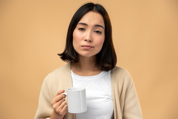 La donna assonnata stanca che tiene una tazza di caffè, guardando la telecamera con un'espressione stanca, non può svegliarsi la mattina e andare al lavoro. Fondo beige isolato