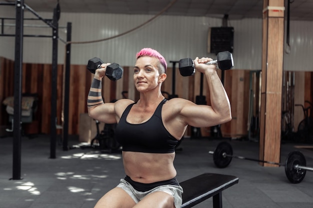 La donna atletica forte allena i muscoli della spalla con i manubri nelle sue mani mentre è seduta su una panchina in una palestra trasversale. Bodybuilding e Fitness